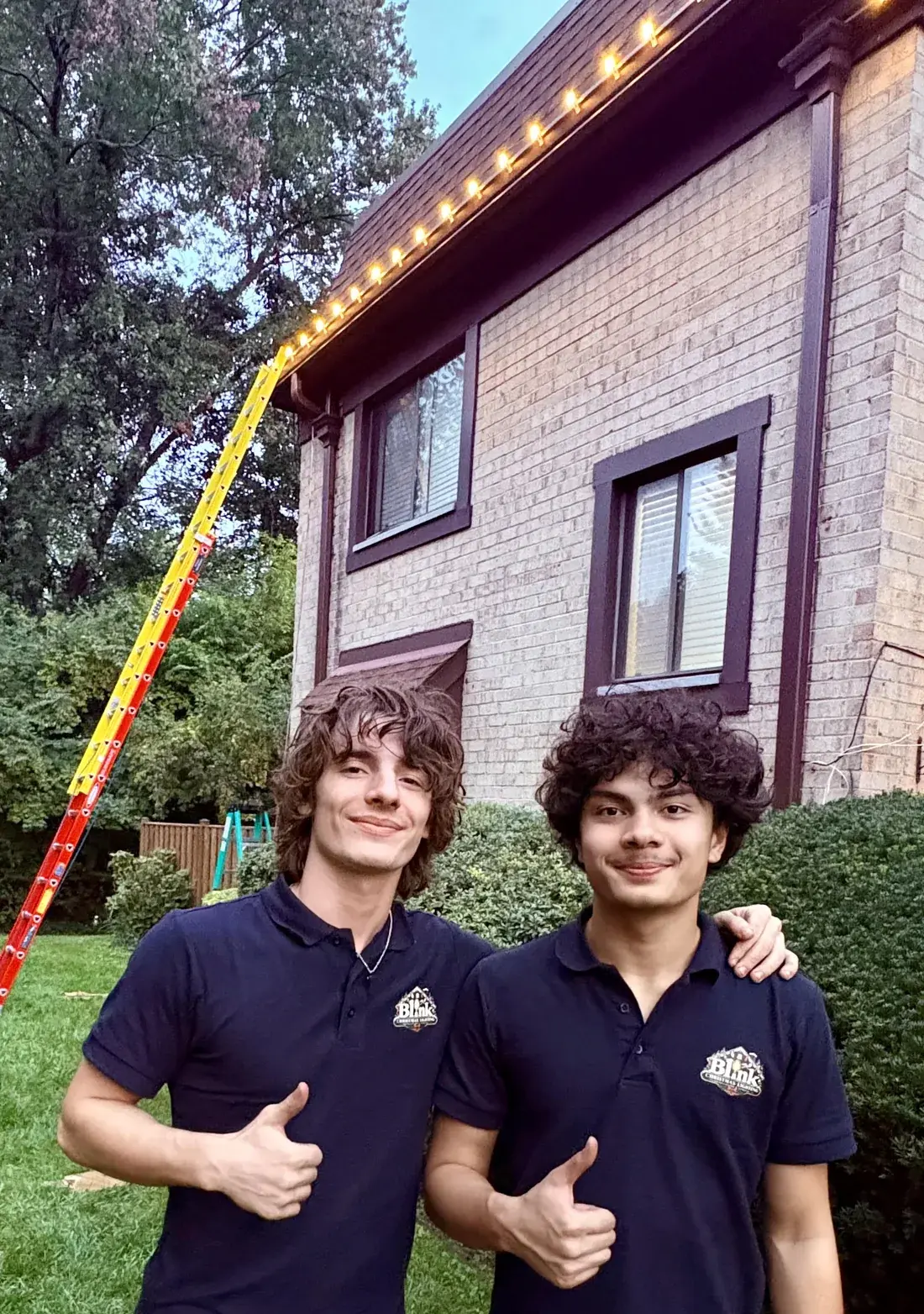 Two members of the Blink crew in Blink polos, giving a thumbs up in front of a brick house with warm white C9s along the roofline and a ladder still up.