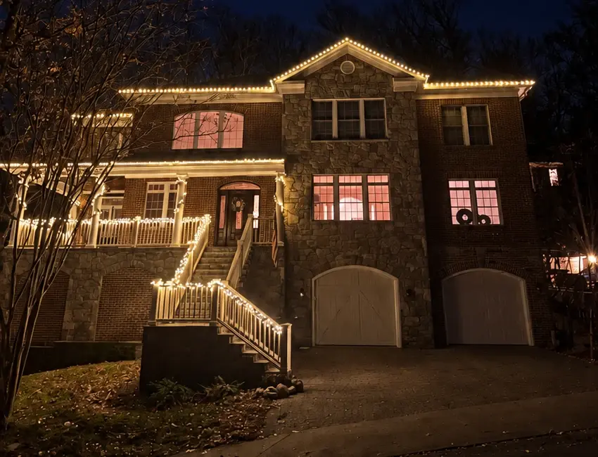 A brick and stone house lit by Blink with warm white lights on the rooflines, porch and front stairs.
