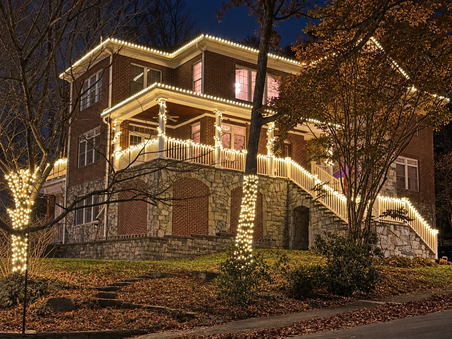 Three-story brick and stone house with warm white lights on every roofline, the porch railings and staircase, and two wrapped trees.